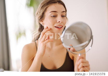 Young woman with problem skin looking into mirror. Inflammation on face, problem skin. Beauty care. 141915378