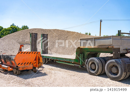 A bustling construction site featuring an orange piece of heavy machinery and a gravel pile, viewed from the side of a dump truck. The scene is set in an industrial environment with visible. A bustling construction site featuring an orange piece of heavy machinery and a gravel pile, viewed from the side of a dump truck. The scene is set in an industrial environment with visible. 141915499