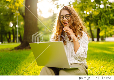 Young woman in glasses and with laptop working at sunset in park on green meadow. Lifestyle concept. 141915568