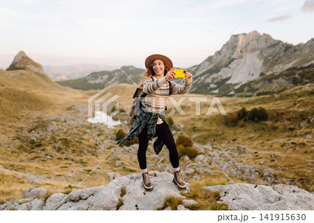 Woman stands on rocky terrain, phone in hand, taking picture of breathtaking landscape. Blogging. 141915630