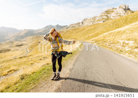 Woman with backpack walking along road with outstretched hand catching car with mountain landscape. Woman with backpack walking along road with outstretched hand catching car with mountain landscape. 141915651