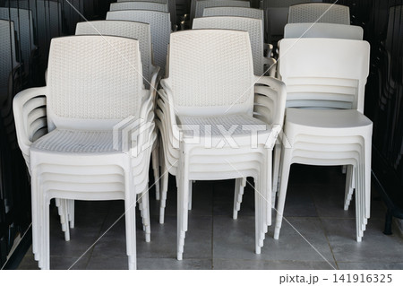 White plastic chairs stacked outdoors at a restaurant or resort, empty seating arrangement with repetitive pattern and modern Mediterranean summer atmosphere White plastic chairs stacked outdoors at a restaurant or resort, empty seating arrangement with repetitive pattern and modern Mediterranean summer atmosphere 141916325