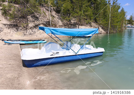 White pedal boat with blue canopy moored at a sandy lakeshore, surrounded by forest and calm green water on a sunny summer day. Ideal for travel, vacation and outdoor recreation concepts White pedal boat with blue canopy moored at a sandy lakeshore, surrounded by forest and calm green water on a sunny summer day. Ideal for travel, vacation and outdoor recreation concepts 141916386