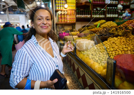 Smiling woman shops at a bustling Mediterranean market stall overflowing with olives, pickles and preserved vegetables, enjoying vibrant colors, local flavors and a friendly food market atmosphere. 141916500