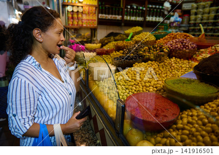 Woman sampling an olive at a bustling market stall surrounded by bowls of olives, pickles, spices and preserved foods, enjoying local flavors and culinary exploration. 141916501