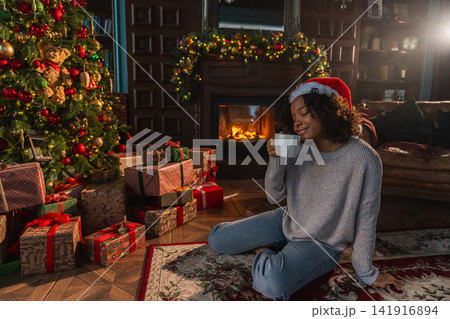 Merry Christmas. African American woman in Santa hat with cup of hot drink coffee tea near Christmas tree in dark interior. Girl in living room with Christmas tree and fireplace. Christmas eve at home Merry Christmas. African American woman in Santa hat with cup of hot drink coffee tea near Christmas tree in dark interior. Girl in living room with Christmas tree and fireplace. Christmas eve at home 141916894