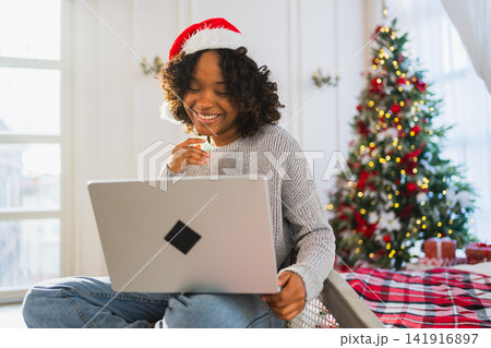 Merry Christmas. African woman in Santa hat having video chat in laptop. Blogger in festive mood recording video in social network. Girl having video call with friends family celebrating Christmas Merry Christmas. African woman in Santa hat having video chat in laptop. Blogger in festive mood recording video in social network. Girl having video call with friends family celebrating Christmas 141916897
