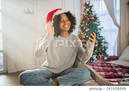 Merry Christmas. African woman in Santa hat having video chat in smartphone. Blogger in festive mood recording video in social network. Girl having video call with friends family celebrating Christmas Merry Christmas. African woman in Santa hat having video chat in smartphone. Blogger in festive mood recording video in social network. Girl having video call with friends family celebrating Christmas 141916899