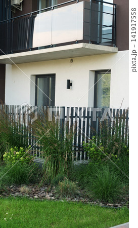 Modern residential building exterior with a glass balcony, white facade, and lush green garden landscaping Modern residential building exterior with a glass balcony, white facade, and lush green garden landscaping 141917558
