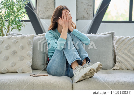 Upset young woman covering face with hands on sofa at home 141918027