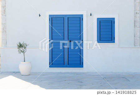 Blue shutters decorate a white Mediterranean house facade with Olea europaea in a pot. Concept of architecture, harmony and coastal lifestyle Blue shutters decorate a white Mediterranean house facade with Olea europaea in a pot. Concept of architecture, harmony and coastal lifestyle 141918825
