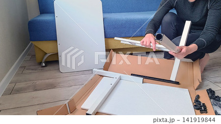 Person assembling white furniture from flat pack box on wooden floor Person assembling white furniture from flat pack box on wooden floor 141919844