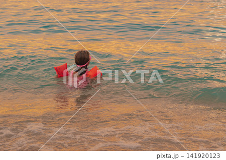 Rear view of little girl swimming in calm sea water with orange inflatable armbands in pink swimsuit during warm golden sunset light childhood vacation photo 141920123