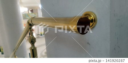Focus on a polished golden handrail end cap, mounted on a marble wall. Leads to an ornate baluster and a long grand staircase descending with marble steps into a lobby. Elegant, classical interior. Focus on a polished golden handrail end cap, mounted on a marble wall. Leads to an ornate baluster and a long grand staircase descending with marble steps into a lobby. Elegant, classical interior. 141920287