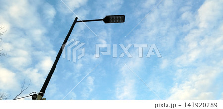 A modern, black LED street lamp against a bright blue, cloud-filled daytime sky. A CCTV camera and junction box are at the pole's base. Shot from a low angle. A modern, black LED street lamp against a bright blue, cloud-filled daytime sky. A CCTV camera and junction box are at the pole's base. Shot from a low angle. 141920289