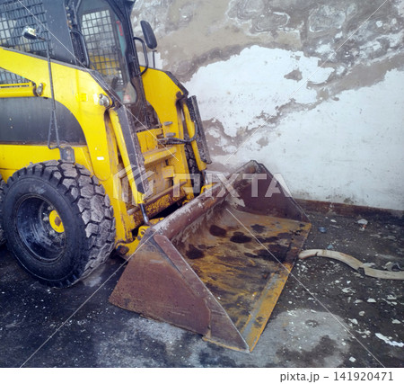A close-up view of a worn, yellow skid-steer loader with its rusty metal front bucket resting on a dirty ground surface next to a peeling, weathered concrete wall. 141920471