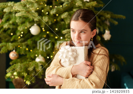 Teen girl hugging Christmas present beside decorated holiday tree 141920935