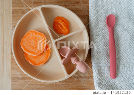 Top view flat lay of a beige silicone baby plate filled with steamed sweet potato slices, teether, pink spoon, and towel on wooden background. Ideal for baby weaning and child care themes 141922129