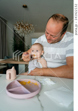 Loving dad holding his baby on his lap at home, feeding fruit puree with modern silicone dishware. Perfect for family relationship concepts, interior lifestyle, and baby nutrition Loving dad holding his baby on his lap at home, feeding fruit puree with modern silicone dishware. Perfect for family relationship concepts, interior lifestyle, and baby nutrition 141922132
