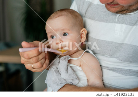 Close-up portrait of an infant baby eating pureed food from a spoon held by father. Ideal for complementary feeding, baby nutrition campaigns, pediatric articles, and child care stock 141922133