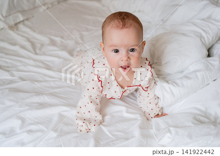 Cute infant lying on tummy and looking up on a white duvet sheets in a bright bedroom. Perfect stock image for baby care products, pediatric health, early childhood, and maternal care stock 141922442