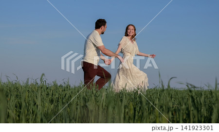 Happy couple holding hands while running through tall green meadow under clear blue sky, smiling woman in flowing white dress and partner in light shirt enjoying warm summer romance 141923301