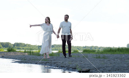 Romantic couple walking hand in hand along grassy riverbank with woman in flowing white dress gesturing beside calm water, with couple, romantic, walking, hand detail Romantic couple walking hand in hand along grassy riverbank with woman in flowing white dress gesturing beside calm water, with couple, romantic, walking, hand detail 141923302