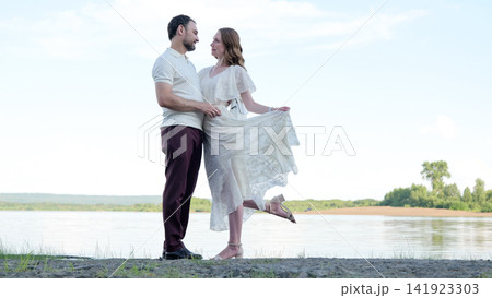 Romantic couple embracing on lakeshore, woman lifting lace dress while smiling at partner beside calm water, pale sky, distant trees and soft daylight shaping serene outdoor mood 141923303