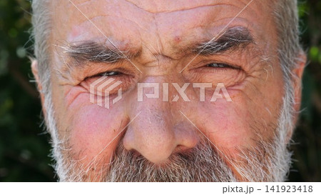 Blue eyes of elderly male blinking and looking into camera outdoor. Wrinkled face of senior human with a sad sight. Upset facial expression of pensioner. Close up Blue eyes of elderly male blinking and looking into camera outdoor. Wrinkled face of senior human with a sad sight. Upset facial expression of pensioner. Close up 141923418