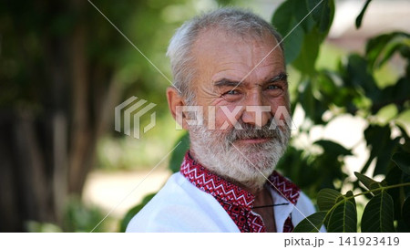 Portrait of old ukrainian man in vyshyvanka near green walnut tree of summer garden. Senior smiling male wearing an embroidered shirt looking into camera outdoor. National identity and patriotism Portrait of old ukrainian man in vyshyvanka near green walnut tree of summer garden. Senior smiling male wearing an embroidered shirt looking into camera outdoor. National identity and patriotism 141923419