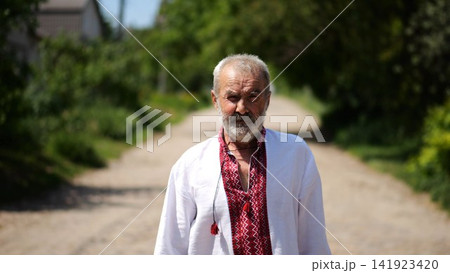 Elderly man wearing an embroidered shirt walking through rural road at sunny summer say. Senior male in traditional vyshyvanka going along country trail. Concept of national identity and patriotism Elderly man wearing an embroidered shirt walking through rural road at sunny summer say. Senior male in traditional vyshyvanka going along country trail. Concept of national identity and patriotism 141923420