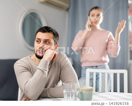 Sad man sitting at table at home, wife talking on phone Sad man sitting at table at home, wife talking on phone 141926158