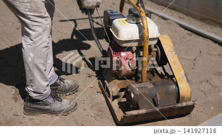 Worker operates heavy plate compactor on dry soil at construction site, flattening ground for foundation preparation, with worn boots, engine unit, metal frame, and strong Worker operates heavy plate compactor on dry soil at construction site, flattening ground for foundation preparation, with worn boots, engine unit, metal frame, and strong 141927118
