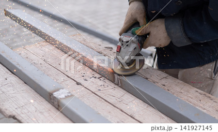 Close view of worker using handheld angle grinder on metal beam, creating shower of bright sparks over rough wooden support boards in industrial construction setting with focused manual effort Close view of worker using handheld angle grinder on metal beam, creating shower of bright sparks over rough wooden support boards in industrial construction setting with focused manual effort 141927145