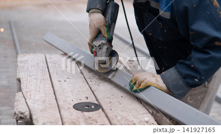 Close view of metal fabrication work with gloved worker guiding grinder along steel beam on rough wooden surface, sending sparks across industrial workspace during careful repair and construction Close view of metal fabrication work with gloved worker guiding grinder along steel beam on rough wooden surface, sending sparks across industrial workspace during careful repair and construction 141927165