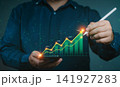 Businessman using stylus pen on smartphone displaying virtual rising stock market graph with bar charts, financial analysis and investment growth concept. 141927283