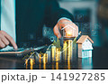 Businessman touching virtual house icon on growing gold coin stacks with rising financial graph and house model, property investment concept. 141927285