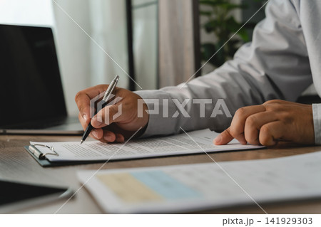 Business person reviewing a printed document with a pen on a clipboard beside a laptop, showing an office document checking process for paperwork verification and administrative review. Business person reviewing a printed document with a pen on a clipboard beside a laptop, showing an office document checking process for paperwork verification and administrative review. 141929303