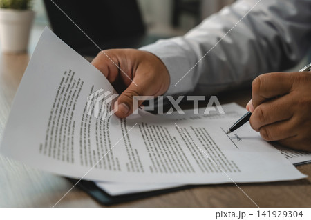 Close-up of hands reviewing printed documents and signing on a marked line with a pen at a desk, showing a contract approval process and business agreement workflow in a modern office. 141929304