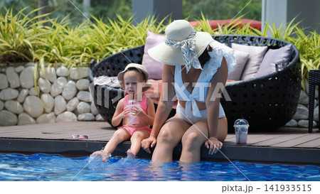 Mother in straw hat and white swimsuit with little daughter in pink swimsuit splashing feet in pool water while daughter drinks a strawberry cocktail. Playful poolside concept Mother in straw hat and white swimsuit with little daughter in pink swimsuit splashing feet in pool water while daughter drinks a strawberry cocktail. Playful poolside concept 141933515