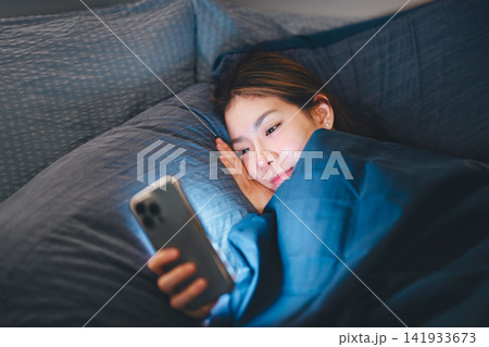 Asian woman lying on bed relaxing at night bedroom scrolling on phone for social media dating online Asian woman lying on bed relaxing at night bedroom scrolling on phone for social media dating online 141933673