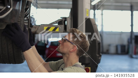 Man repairs car in garage. Car is above person. Employee evaluates technical condition of machine. 141935528