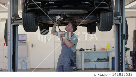Car mechanic is working under a vehicle on a lift in service. Repairman is using ratchet. 141935532