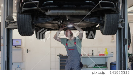 Car service - mechanic check out automobile parts while working with lamp. Auto mechanic working underneath car lifting machine at the garage. Car service - mechanic check out automobile parts while working with lamp. Auto mechanic working underneath car lifting machine at the garage. 141935533