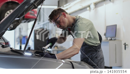 Professional mechanic working on a vehicle in a car service. Engine specialist fixing the motor. Repairman is wearing gloves and using a ratchet. Professional mechanic working on a vehicle in a car service. Engine specialist fixing the motor. Repairman is wearing gloves and using a ratchet. 141935554
