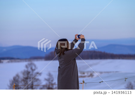 黄昏時の雪原をスマホで撮影する後ろ姿の女性|北海道の冬景色を記録する旅の一コマ 黄昏時の雪原をスマホで撮影する後ろ姿の女性|北海道の冬景色を記録する旅の一コマ 141936113