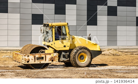 A yellow roller compacts dirt on a construction site next to a building with a gray and black facade. Workers are present in the background 141936414