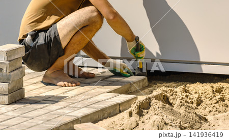 A person works on laying tiles on a ground surface using a trowel. The area has sand and building materials close to a white wall during the day A person works on laying tiles on a ground surface using a trowel. The area has sand and building materials close to a white wall during the day 141936419