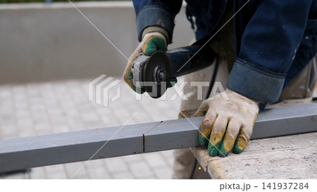 Close view shows worker cutting gray metal beam with angle grinder, wearing protective gloves and work jacket while holding material steady on bench during industrial fabrication and repair task 141937284