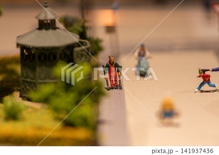 Miniature street diorama with worker beside gazebo and blurred passing figures Miniature street diorama with worker beside gazebo and blurred passing figures 141937436